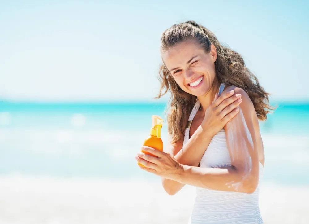 A smiling woman applying sunscreen onto her arms while at the beach